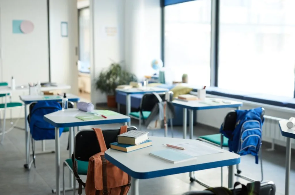 Zdjęcie przedstawiające salę szkolną z biurkami, zeszytami i długopisami